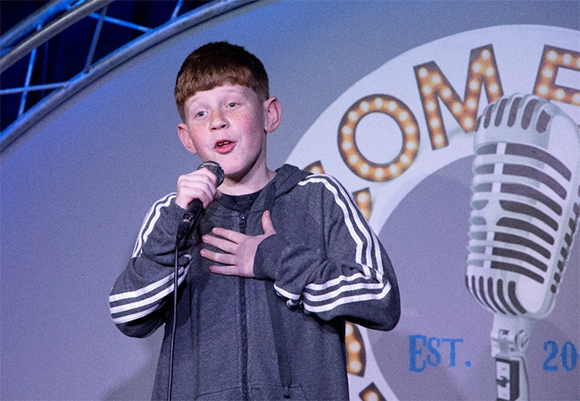 Young boy holding a mic on stage telling a joke