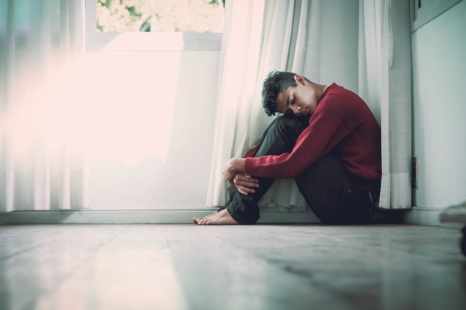 Teenage boy crouched in the corner of a room wearing a red jumper and jeans