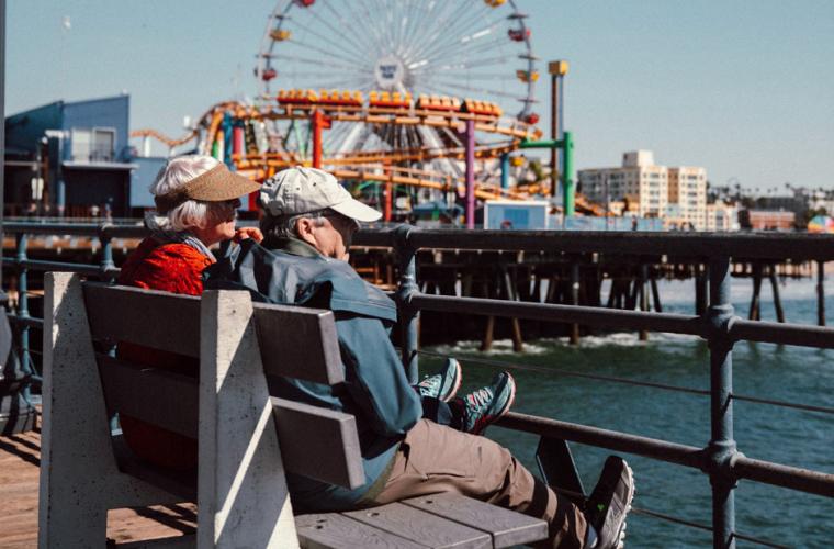 Elderly couple sitting on a bench beside a pier with a Ferris wheel in the background