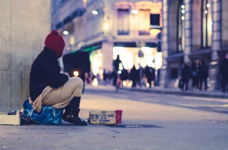 homeless person sitting in street with red hat and a cardboard sign