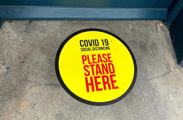 A bright yellow, circular sticker on the ground with the words "Covid-19 social distancing, please stand here" in black and red